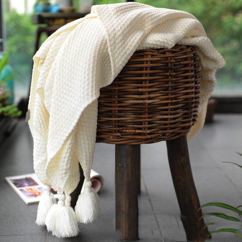 White textured blanket with tassels draped over a wicker stool indoors.