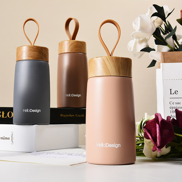 Three insulated drink containers with wooden lids on a surface with books and flowers.