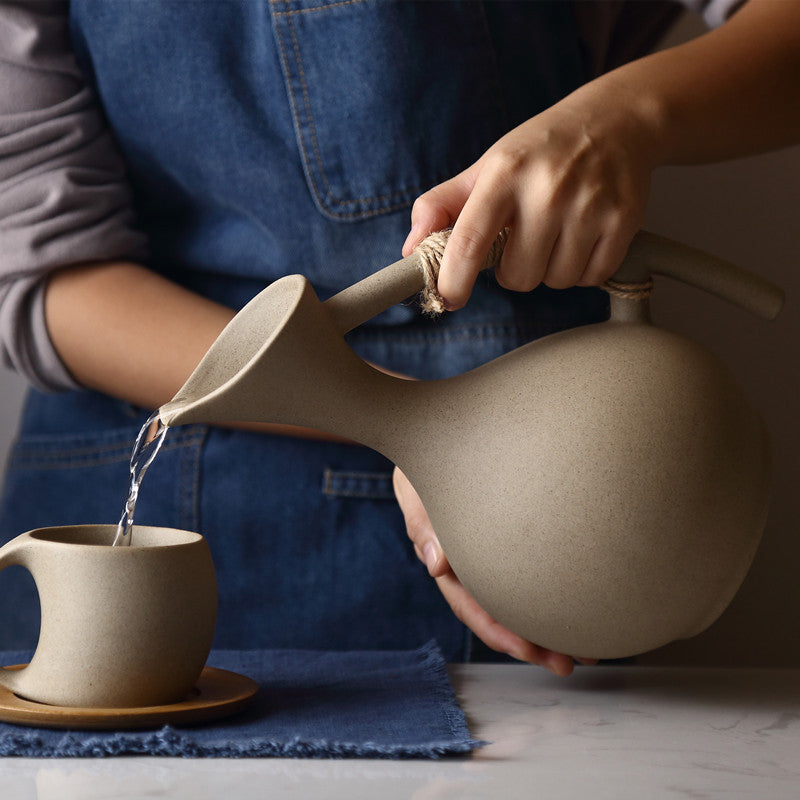 Person pouring water from a ceramic pitcher into a cup on a table.