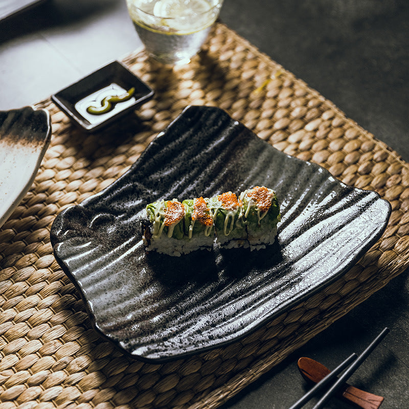 Sushi roll on a black textured plate with a glass of water and wasabi in the background.