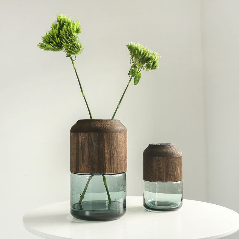 Two glass vases with wooden accents holding green plants on a white surface.