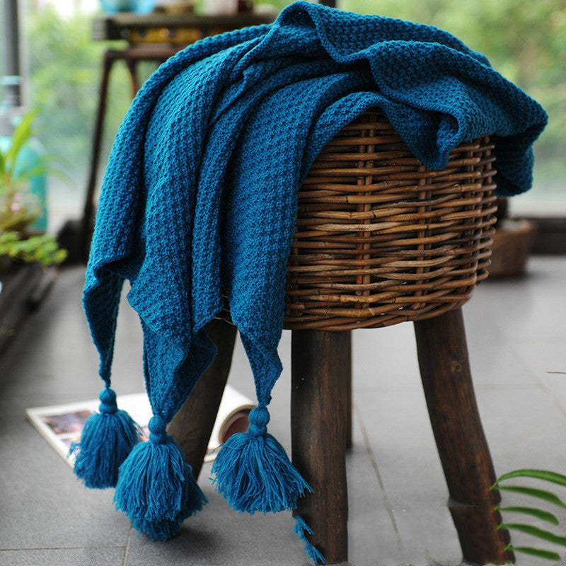 Blue knitted blanket with tassels draped over a wicker stool indoors.