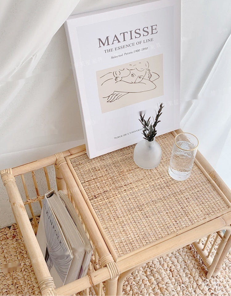 Wicker side table with a glass of water, plant, and magazine against a white wall with 'Matisse' poster.