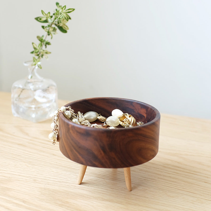 Wooden jewelry bowl with jewelry on a light wooden surface, with a vase of greenery in the background.
