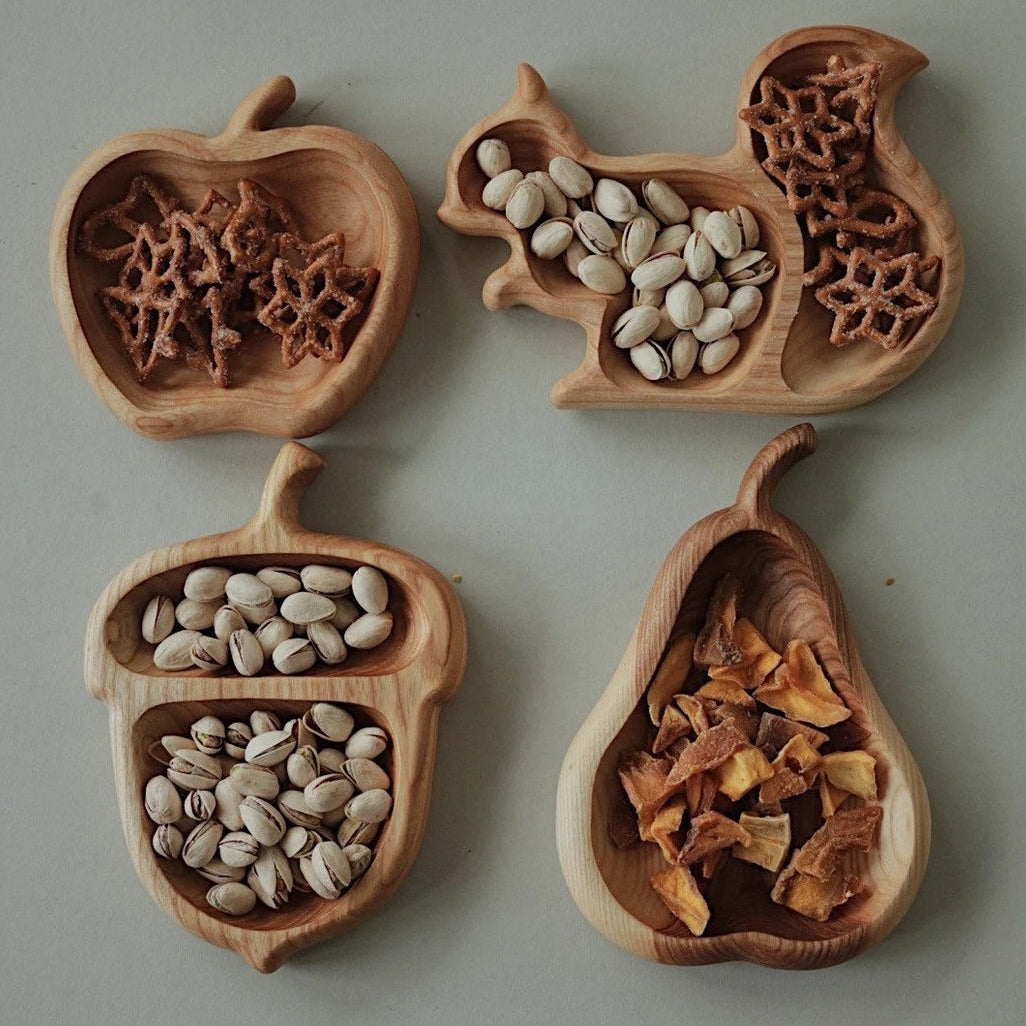Four wooden bowls shaped like an apple, squirrel, acorn, and pear filled with various seeds and nuts on a light gray background.